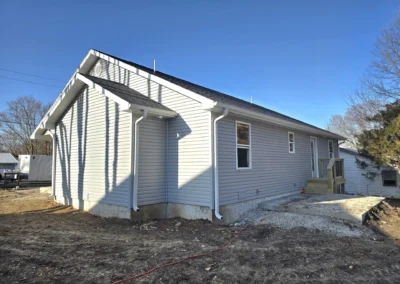 Side view of a newly built light gray house with a wooden deck, surrounded by a cleared ground and under a clear blue sky.